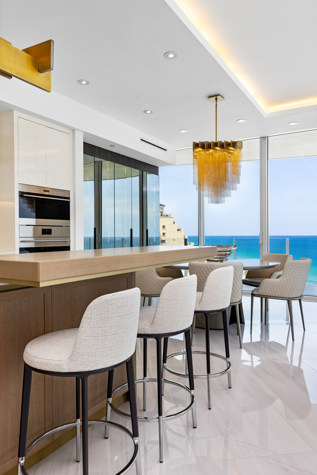 This interior shot showcases a modern kitchen and dining area with a luxurious feel. The kitchen features a breakfast bar with stylish bar stools, while the dining area includes a round table with upholstered chairs, all set against a backdrop of floor-to-ceiling windows offering ocean views. A statement chandelier hangs above the dining table, adding to the upscale ambiance.