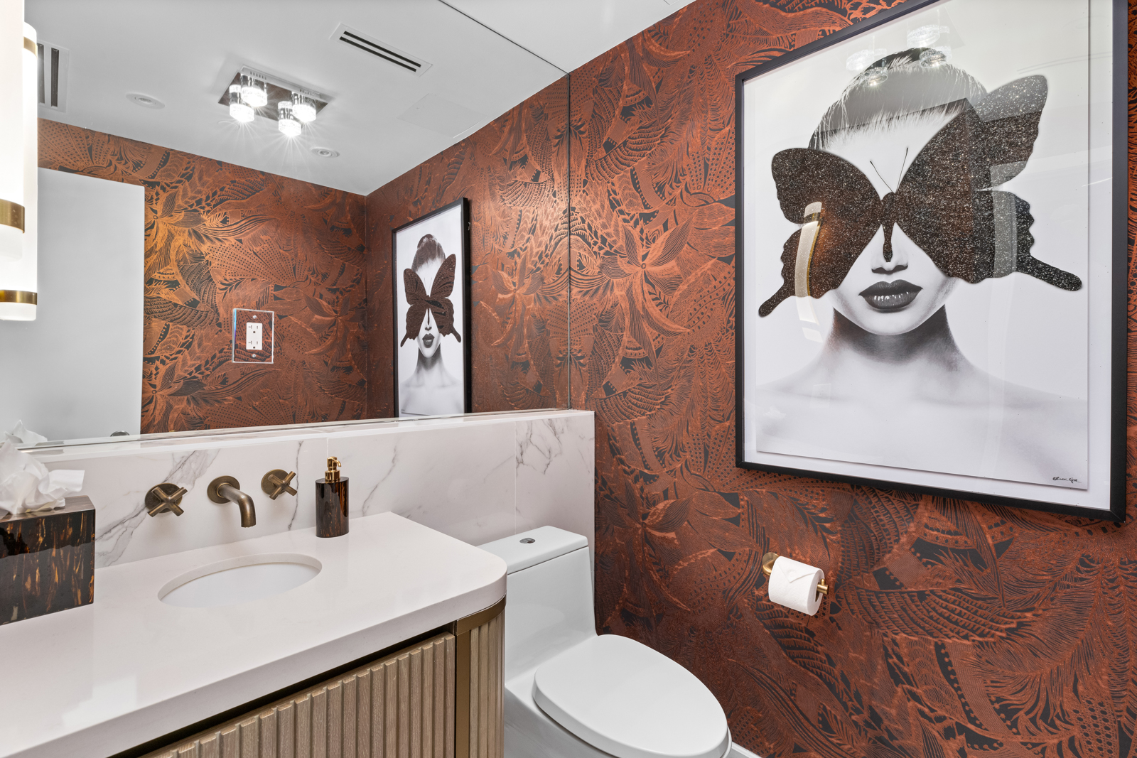 This is a stylish guest bathroom featuring a unique copper-toned patterned wallpaper that adds warmth and texture. The vanity has a light-colored countertop with a modern sink and bronze fixtures, complemented by a sleek, vertically-ribbed cabinet. A framed black and white art piece with a butterfly motif adds a touch of sophistication, while the toilet is neatly tucked beside the vanity.