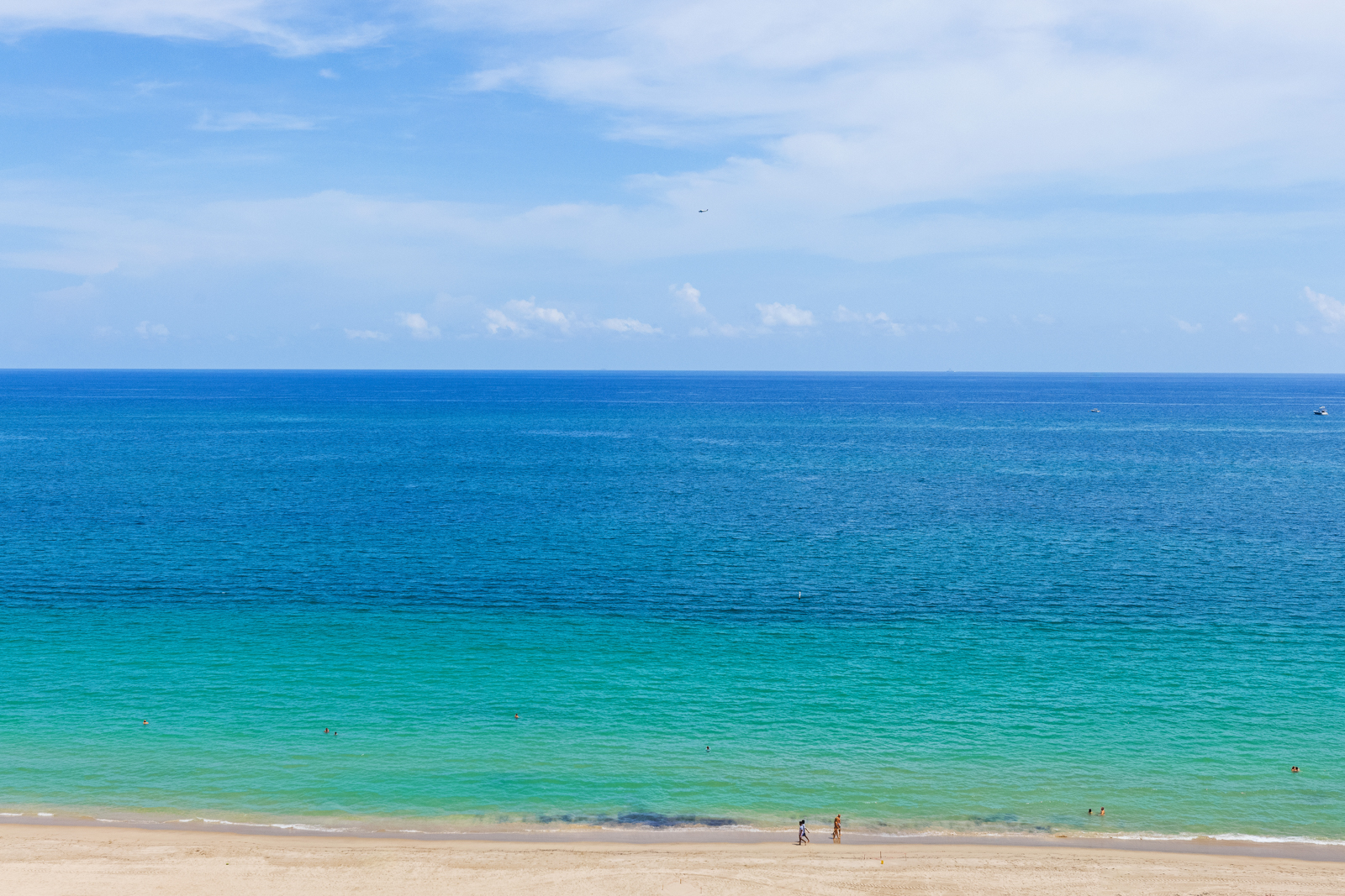 This aerial view showcases a stunning beach scene with a gradient of turquoise to deep blue ocean waters meeting a sandy shoreline. The sky is a clear blue with scattered clouds, creating a serene and inviting atmosphere. The image captures the vastness and beauty of the coastal landscape, perfect for highlighting the location of a beachfront property.