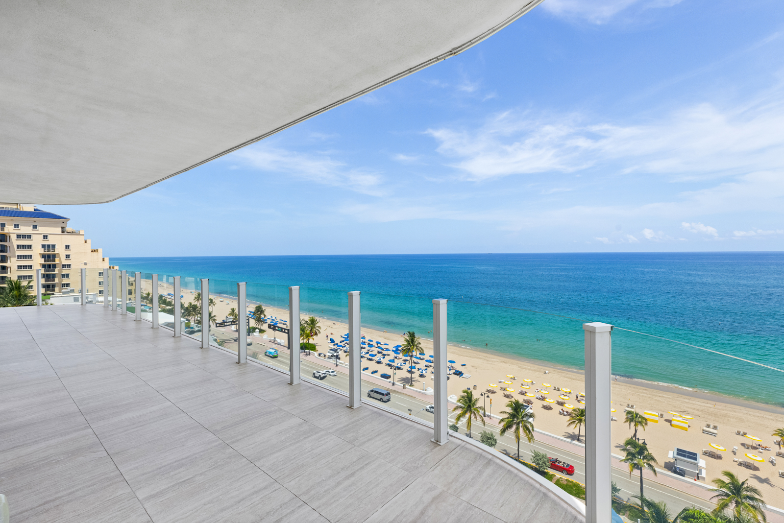 This image showcases a spacious balcony with a stunning ocean view. The balcony features a tiled floor and a modern glass railing, providing an unobstructed view of the beach, palm trees, and the ocean. The scene evokes a sense of luxury and relaxation, perfect for enjoying the coastal scenery.