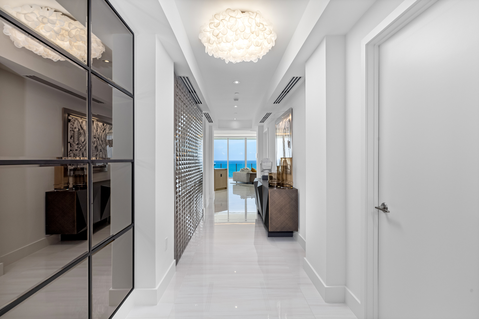 This is a bright and modern hallway featuring glossy white tile flooring and white walls. A decorative mirrored wall and a textured accent wall add visual interest, while a contemporary chandelier provides ambient lighting. The hallway leads to a living area with ocean views, creating a sense of openness and luxury.