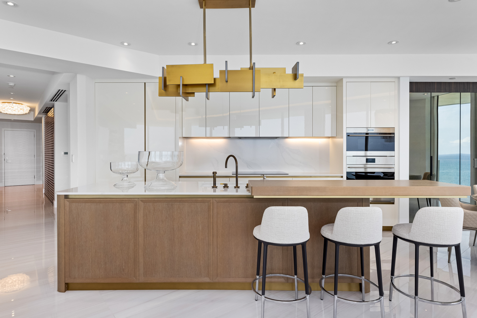 This is a bright and modern kitchen featuring sleek, white cabinetry and a contrasting wood-paneled island with a white countertop. A unique gold chandelier hangs above the island, which is complemented by three stylish bar stools. The kitchen is well-lit and appears to be part of an open-concept living space, with a glimpse of an ocean view through a sliding glass door.