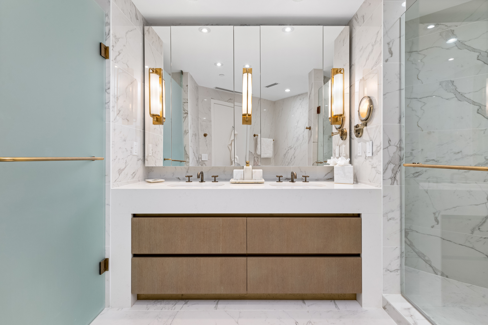 This is a luxurious primary bathroom featuring a double vanity with a white countertop and light wood cabinetry. The walls and floor are covered in marble, and the space is illuminated by modern sconces and recessed lighting. A large mirror spans the length of the vanity, and a glass-enclosed shower is visible on the right.
