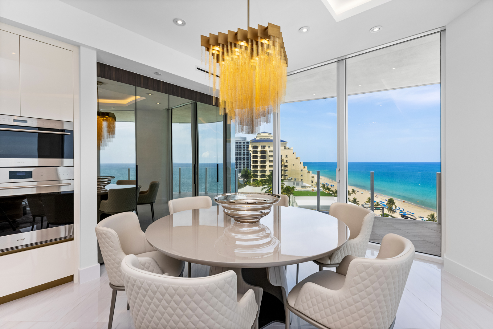 This is a bright and modern dining room featuring a round table with six upholstered chairs. A unique chandelier hangs above the table, and large windows offer stunning ocean views. The room is adjacent to a kitchen area with built-in appliances, creating a seamless and luxurious living space.