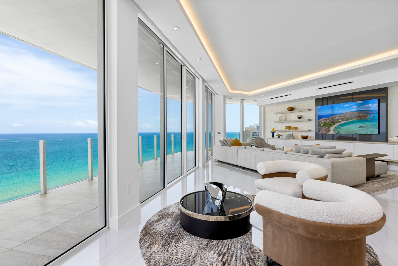 This is a luxurious living room with floor-to-ceiling windows offering stunning ocean views. The room features modern furniture, including a large sectional sofa and unique armchairs, arranged around a stylish coffee table on a patterned rug. The space is bright and airy, with a contemporary design and a large television mounted on the wall.