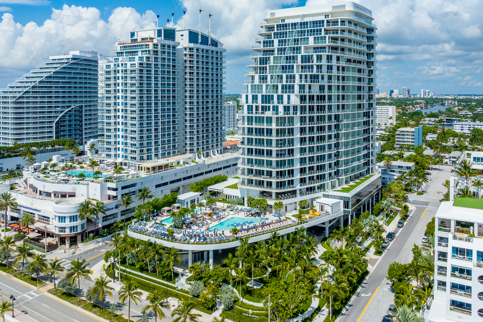 This aerial view showcases a luxurious real estate property featuring modern high-rise buildings with sleek architectural designs. The property includes multiple swimming pools surrounded by lush landscaping, creating a resort-like atmosphere. The scene is set against a backdrop of clear blue skies and distant city views, emphasizing the property's prime location and upscale appeal.