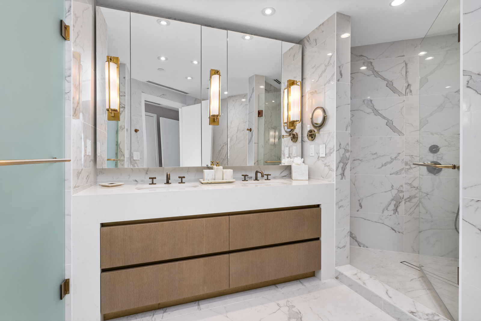 This is a luxurious primary bathroom featuring marble walls and flooring, a double vanity with a white countertop, and modern bronze fixtures. The vanity has wooden drawers and is illuminated by vertical sconces flanking a large mirror. A glass-enclosed shower is visible to the right, completing the elegant and spa-like atmosphere.