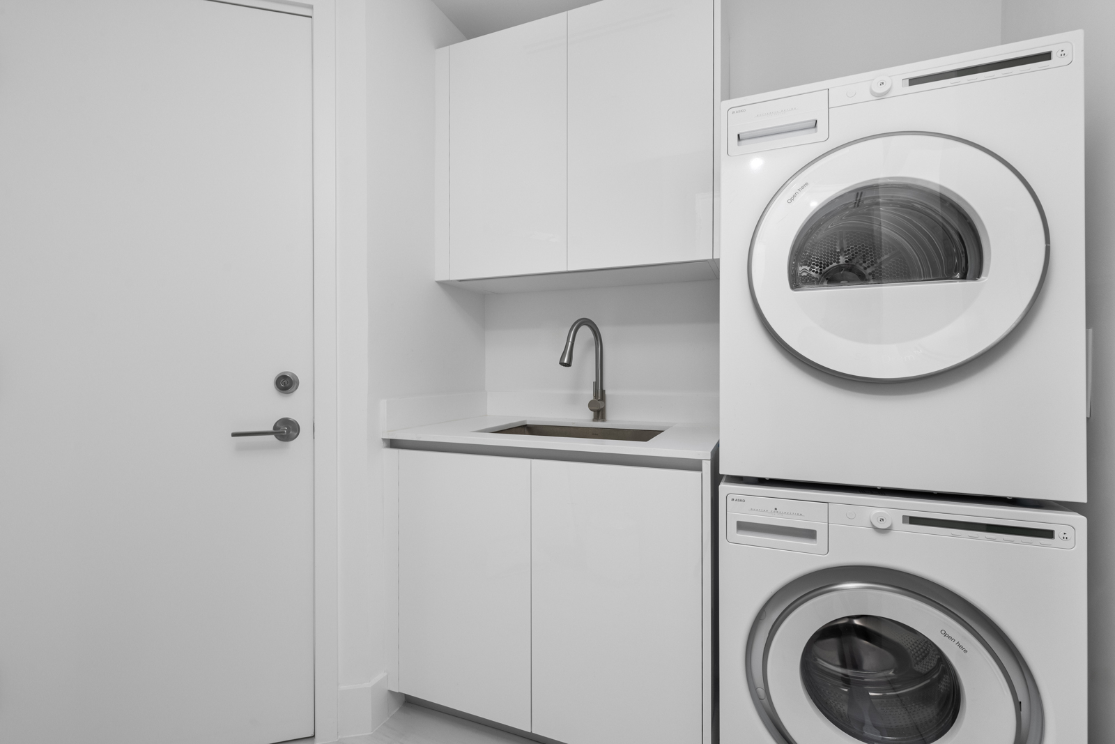 This is a well-lit, modern laundry room featuring a stacked washer and dryer unit in white, complemented by white cabinetry and a sink with a sleek faucet. The room has a clean and minimalist aesthetic, with a focus on functionality and space efficiency. A closed white door is visible on the left.