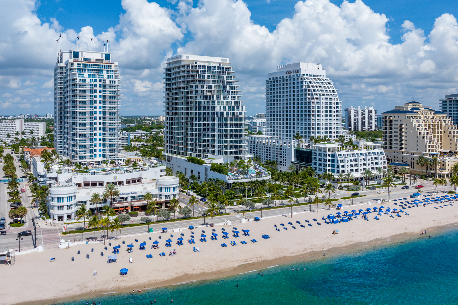 This aerial view showcases a stunning beachfront property featuring multiple high-rise condominium buildings with modern architectural designs. The pristine sandy beach is lined with blue umbrellas and lounge chairs, inviting relaxation and leisure. The turquoise ocean water adds to the luxurious and serene atmosphere, making it an ideal coastal real estate location.