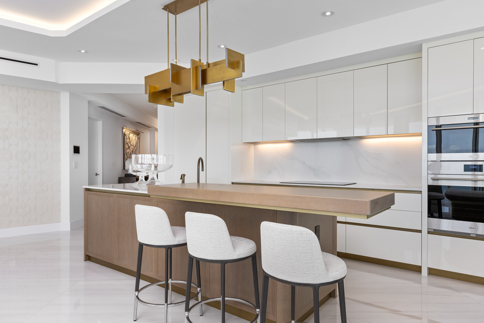 This is a bright, modern kitchen featuring sleek white cabinetry, stainless steel appliances, and a stylish island with a wooden countertop extension. The island is complemented by three bar stools with light-colored upholstery. A unique gold pendant light fixture hangs above the island, adding a touch of elegance to the space.