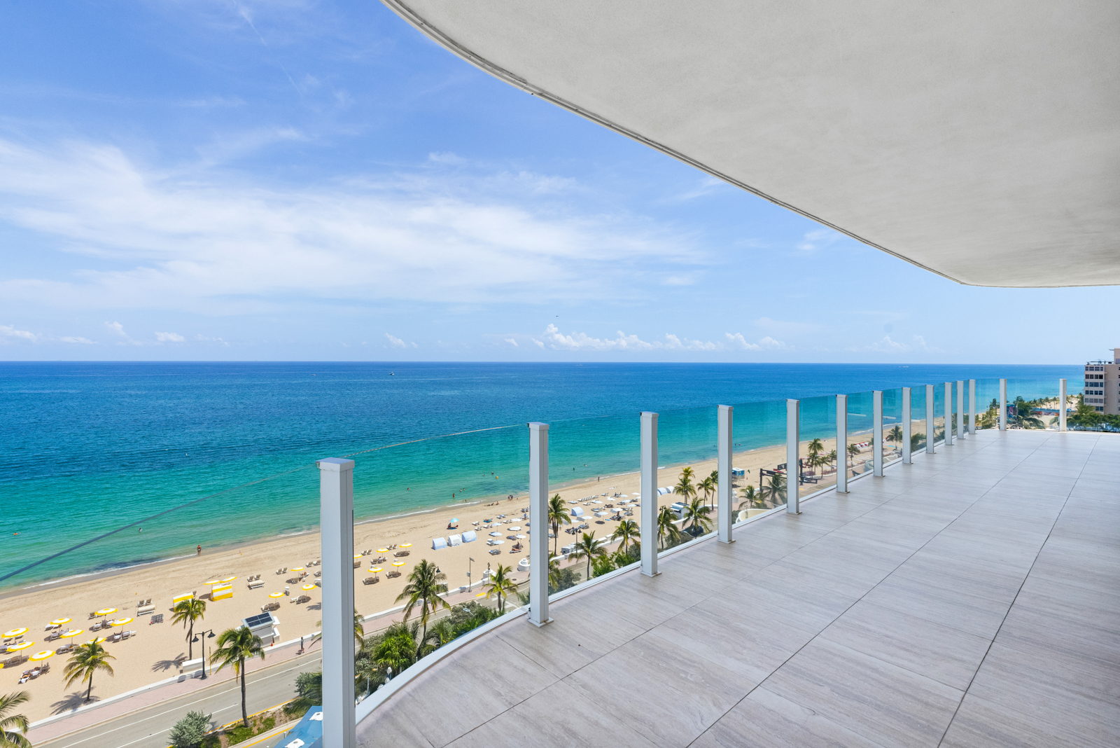 This image showcases a spacious balcony with a stunning ocean view. The balcony features tiled flooring and a modern glass railing system. The view includes a sandy beach with palm trees and clear turquoise water, creating a luxurious and relaxing atmosphere.