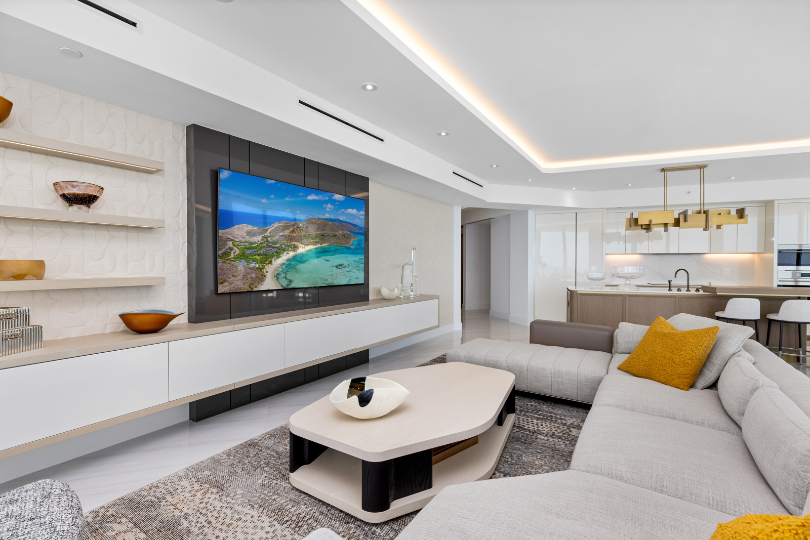 This is a modern living room featuring a large television mounted on a textured wall, complemented by sleek white cabinetry and shelving. A sectional sofa with a mustard-colored pillow sits on a patterned rug, and a unique coffee table adds to the contemporary aesthetic. The open floor plan seamlessly connects to the kitchen area, creating a spacious and inviting atmosphere.