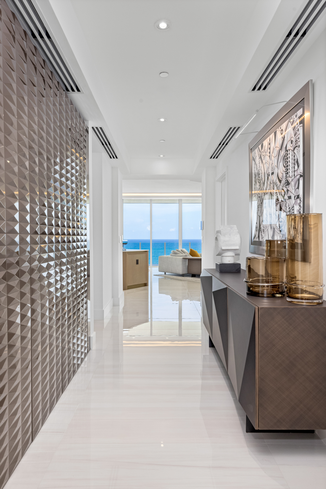 This is a bright and modern hallway featuring glossy white tile flooring and a textured accent wall. A sleek console table with decorative vases and artwork adds a touch of elegance. The hallway leads to a living area with ocean views, creating a sense of openness and luxury.