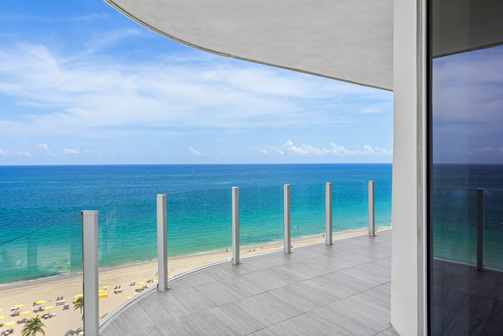This image showcases a luxurious balcony with a stunning ocean view. The balcony features a modern design with gray tiled flooring and a glass railing system, providing an unobstructed view of the turquoise water and sandy beach. The scene evokes a sense of relaxation and high-end coastal living.