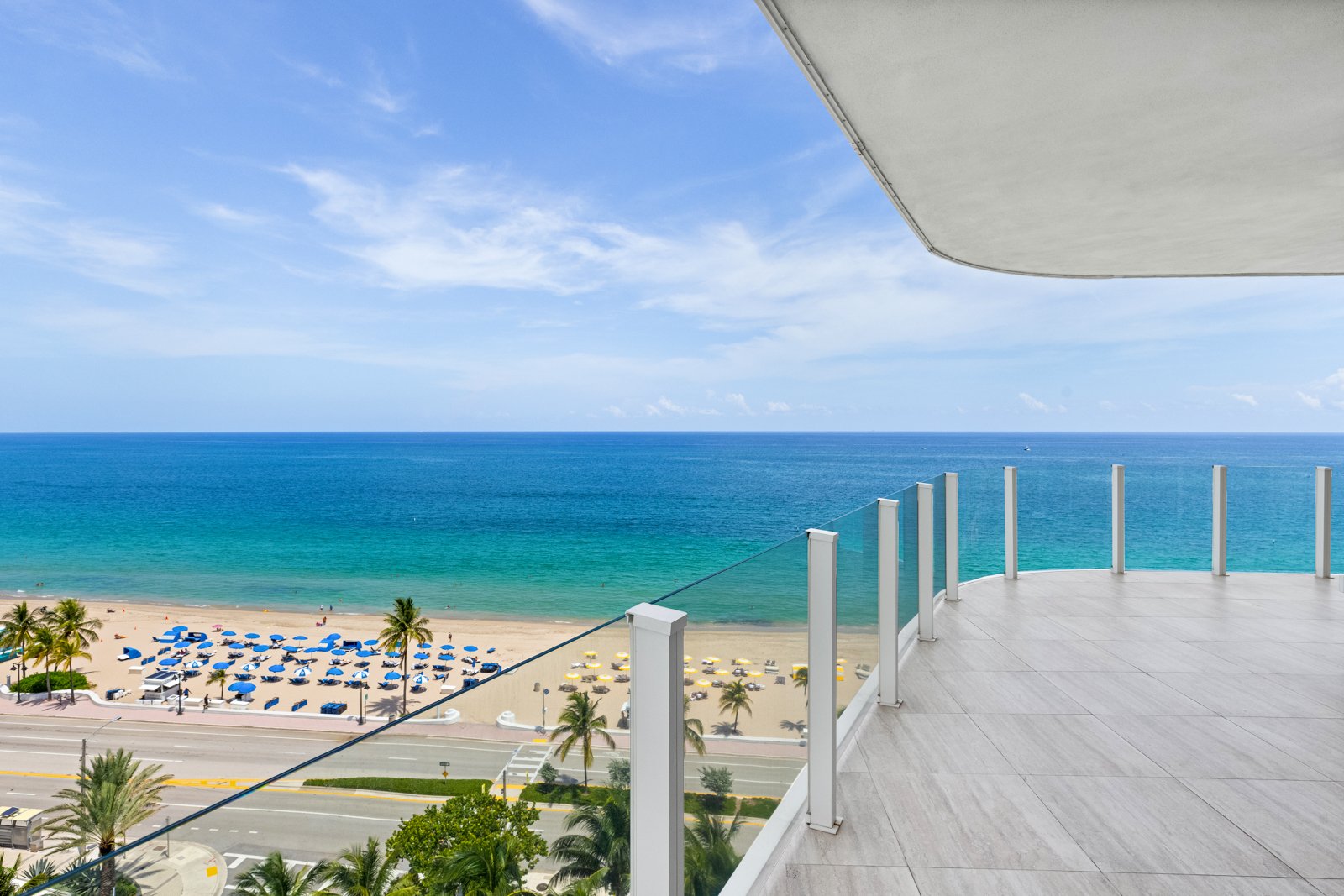 This image showcases a luxurious balcony overlooking a beautiful beach and ocean. The balcony features a modern glass railing and tiled flooring, providing an elegant outdoor living space. The view includes palm trees, beach umbrellas, and the clear blue ocean, creating a serene and inviting atmosphere.