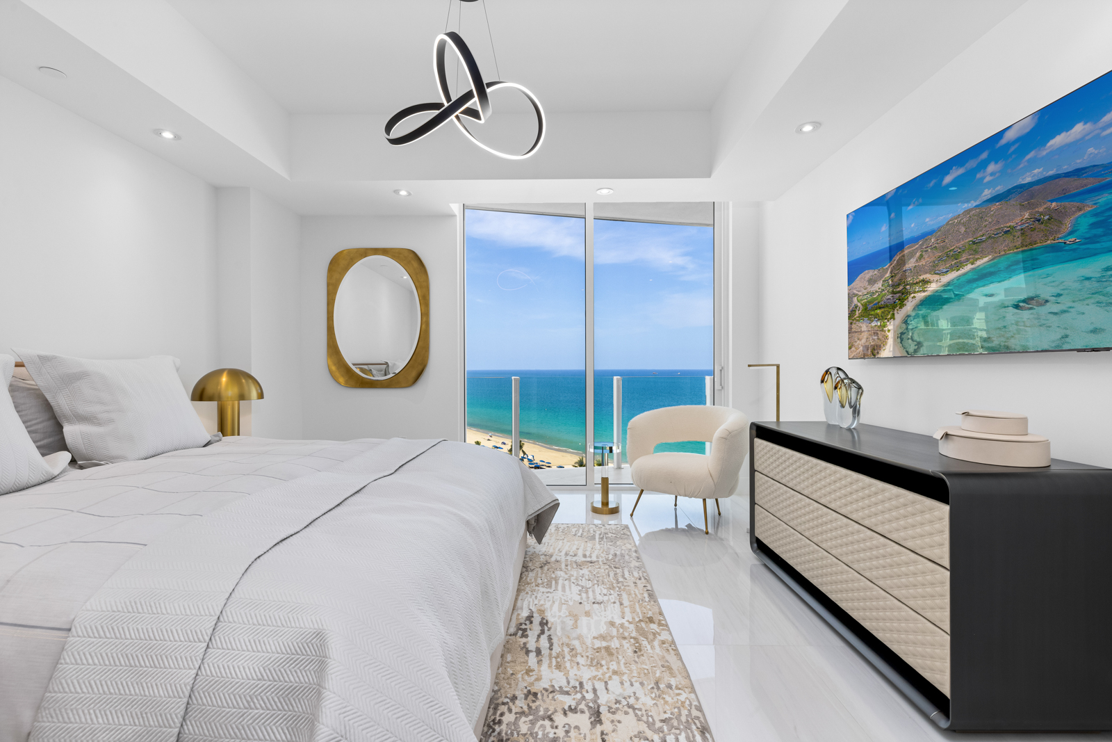 This is a bright and airy primary bedroom featuring a modern design with a luxurious feel. The room boasts a large bed with crisp white linens, a stylish dresser with a flat-screen TV above, and a comfortable chair near a large window offering ocean views. The decor is minimalist yet elegant, with a gold-framed mirror and a unique chandelier adding sophisticated touches.