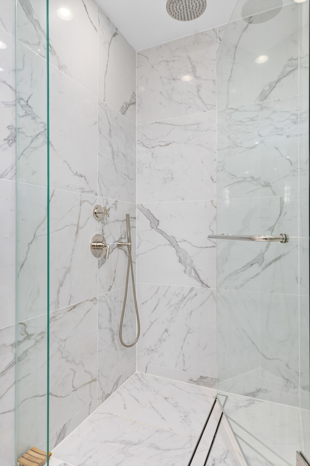 This is a modern primary bathroom featuring white marble walls and flooring, creating a luxurious and clean aesthetic. The shower area is enclosed with glass panels and includes a rainfall showerhead, a handheld shower fixture, and a sleek grab bar. The overall impression is one of high-end design and spa-like tranquility.