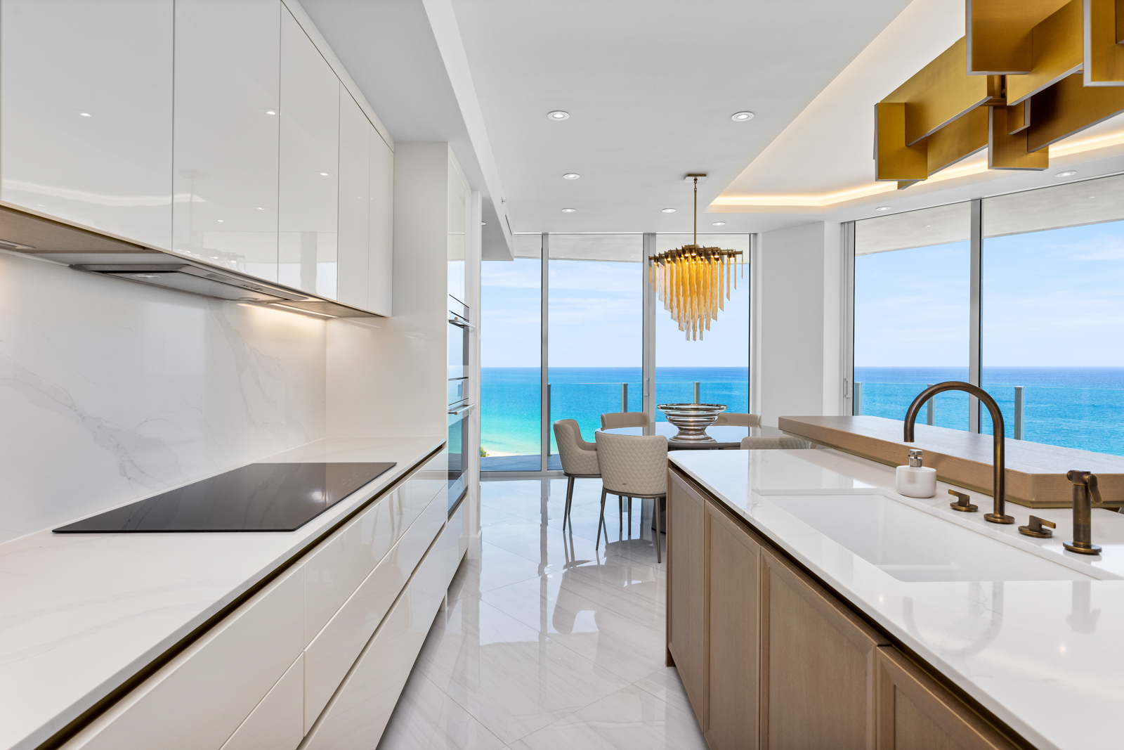 This is a bright and modern kitchen with sleek white cabinetry and a contrasting wood-toned island. The kitchen features high-end appliances and a stunning ocean view through large windows, creating a luxurious and inviting atmosphere. The marble flooring adds to the elegance of the space.