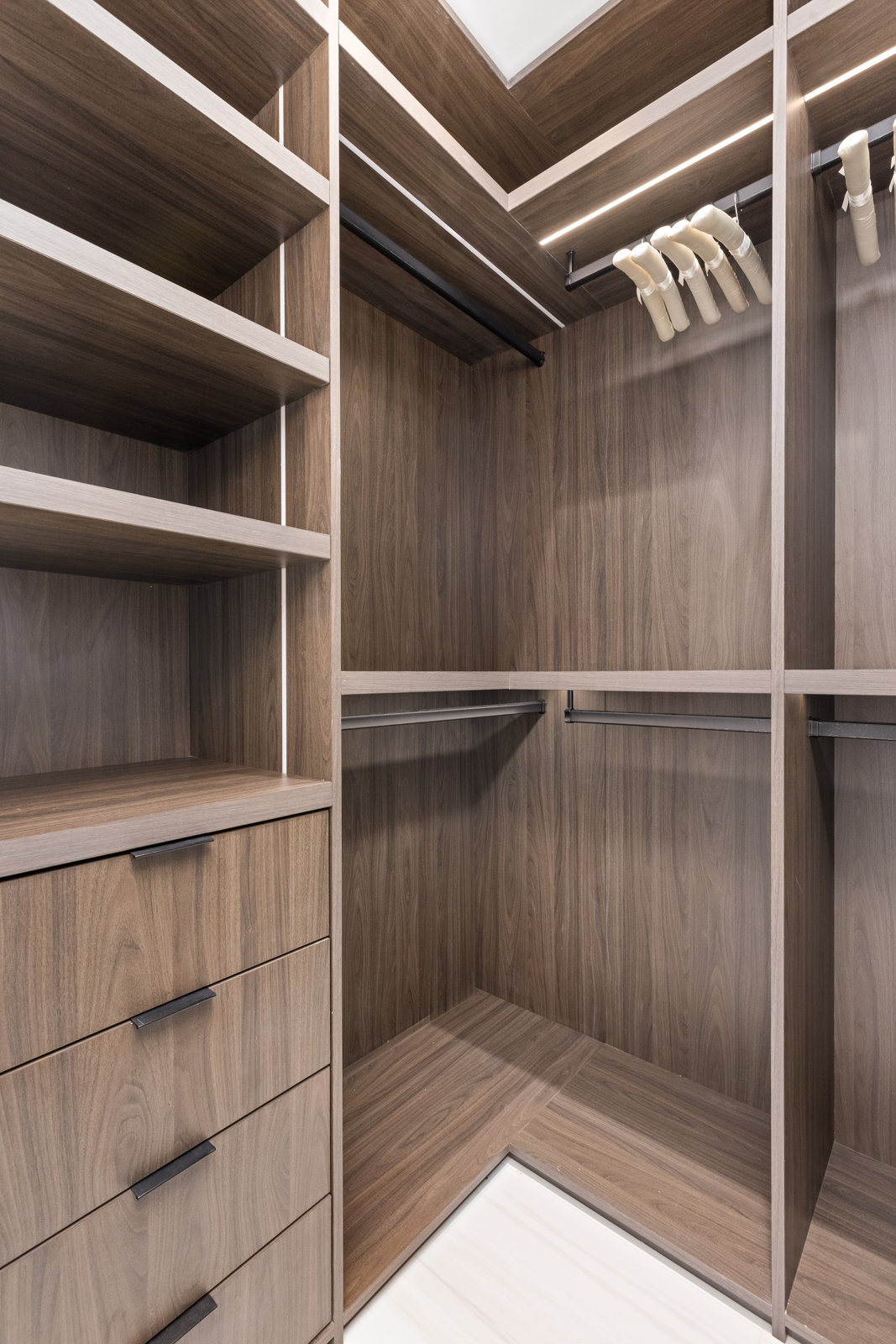 This is a well-organized walk-in closet featuring custom wood shelving, drawers, and hanging rods. The closet is illuminated by integrated lighting, highlighting the clean lines and modern design. The wood grain adds warmth and sophistication to the space, making it an appealing feature for potential buyers.