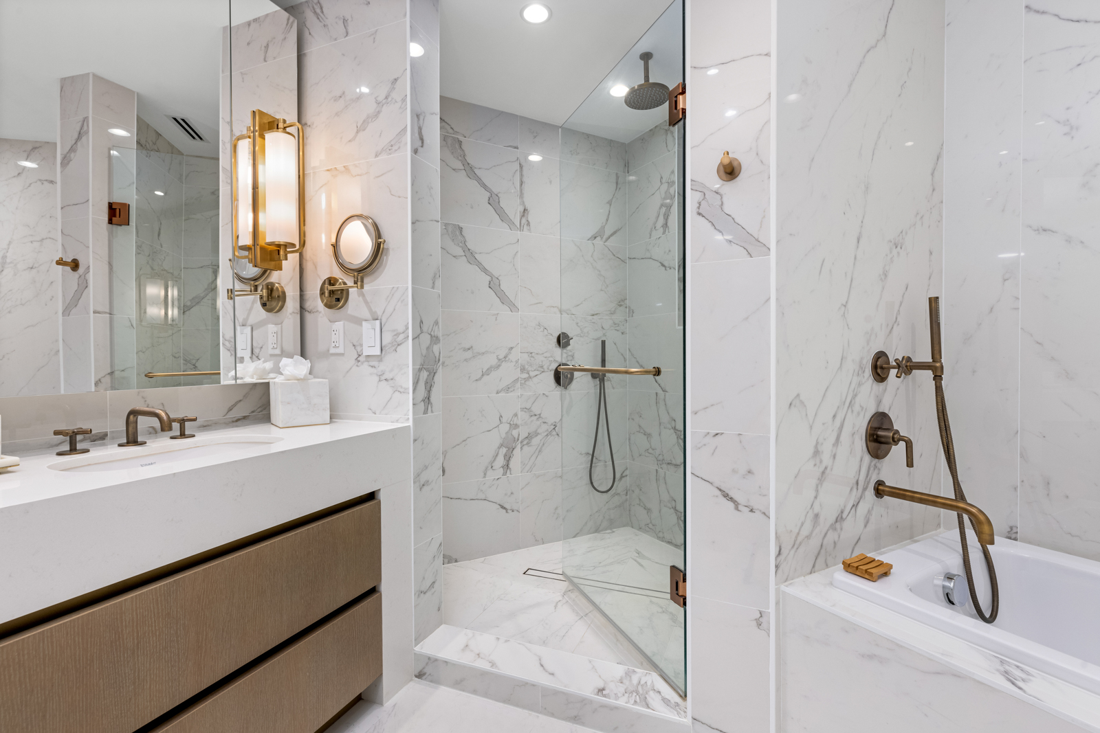 This is a luxurious primary bathroom featuring marble walls and flooring, a glass-enclosed shower with bronze fixtures, and a separate bathtub. The vanity has a white countertop with bronze faucets and wooden drawers, complemented by elegant lighting and a magnifying mirror. The overall impression is one of sophistication and high-end design.