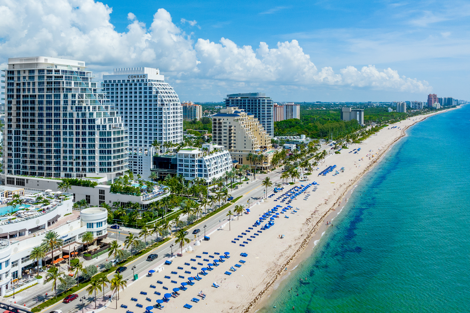 This aerial view showcases a stunning coastal property featuring modern high-rise buildings along a pristine beach. The turquoise ocean contrasts beautifully with the white sand and rows of beach umbrellas. Lush palm trees line the street, adding to the tropical ambiance and highlighting the desirable location.