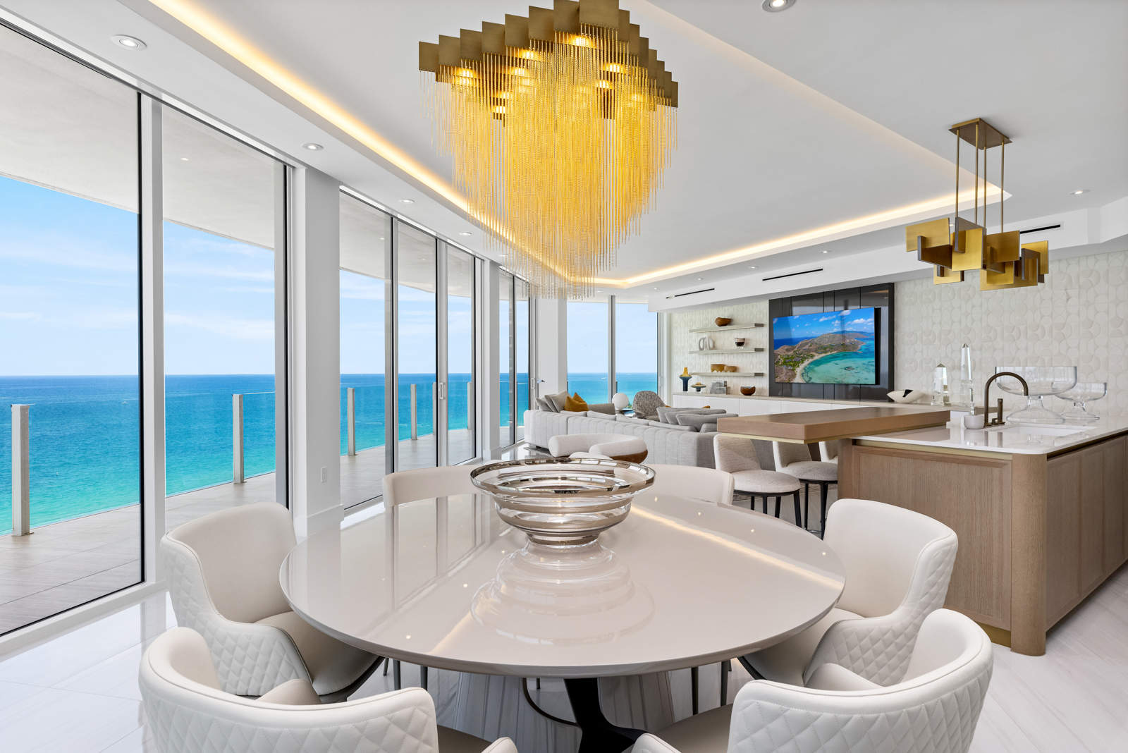This interior shot showcases a luxurious dining area with a round table and plush white chairs, complemented by a modern chandelier. Floor-to-ceiling windows offer stunning ocean views, enhancing the room's bright and airy atmosphere. The open-concept design seamlessly integrates the dining space with the living area and kitchen, creating a cohesive and elegant living environment.