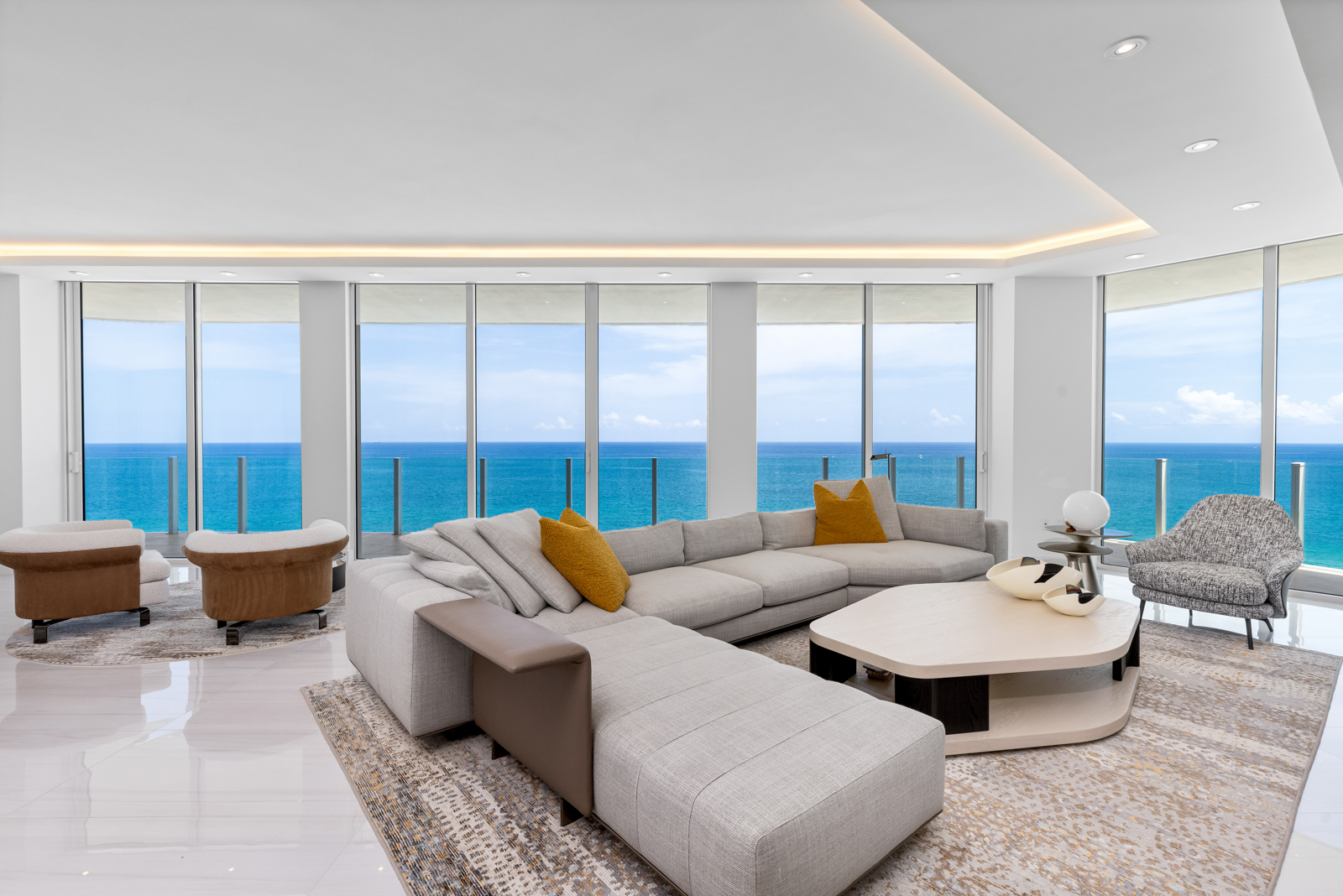 This is a luxurious living room with a modern design, featuring a large sectional sofa with mustard-colored pillows, a unique coffee table, and two stylish armchairs. The room boasts floor-to-ceiling windows offering stunning ocean views, and the white marble floors and recessed lighting contribute to a bright and airy atmosphere. The perspective is from a medium shot, showcasing the entire living room.