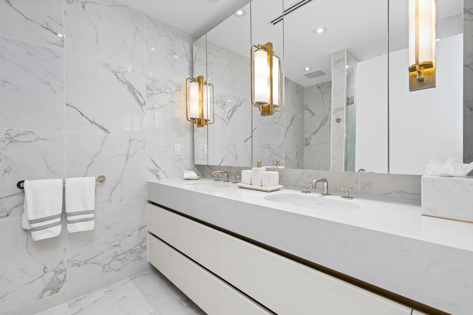 This is a luxurious primary bathroom featuring a double vanity with white countertops and cabinetry, complemented by marble-patterned walls and flooring. Modern gold-accented light fixtures flank the large mirror, adding a touch of elegance. The space is bright, clean, and designed with high-end finishes.