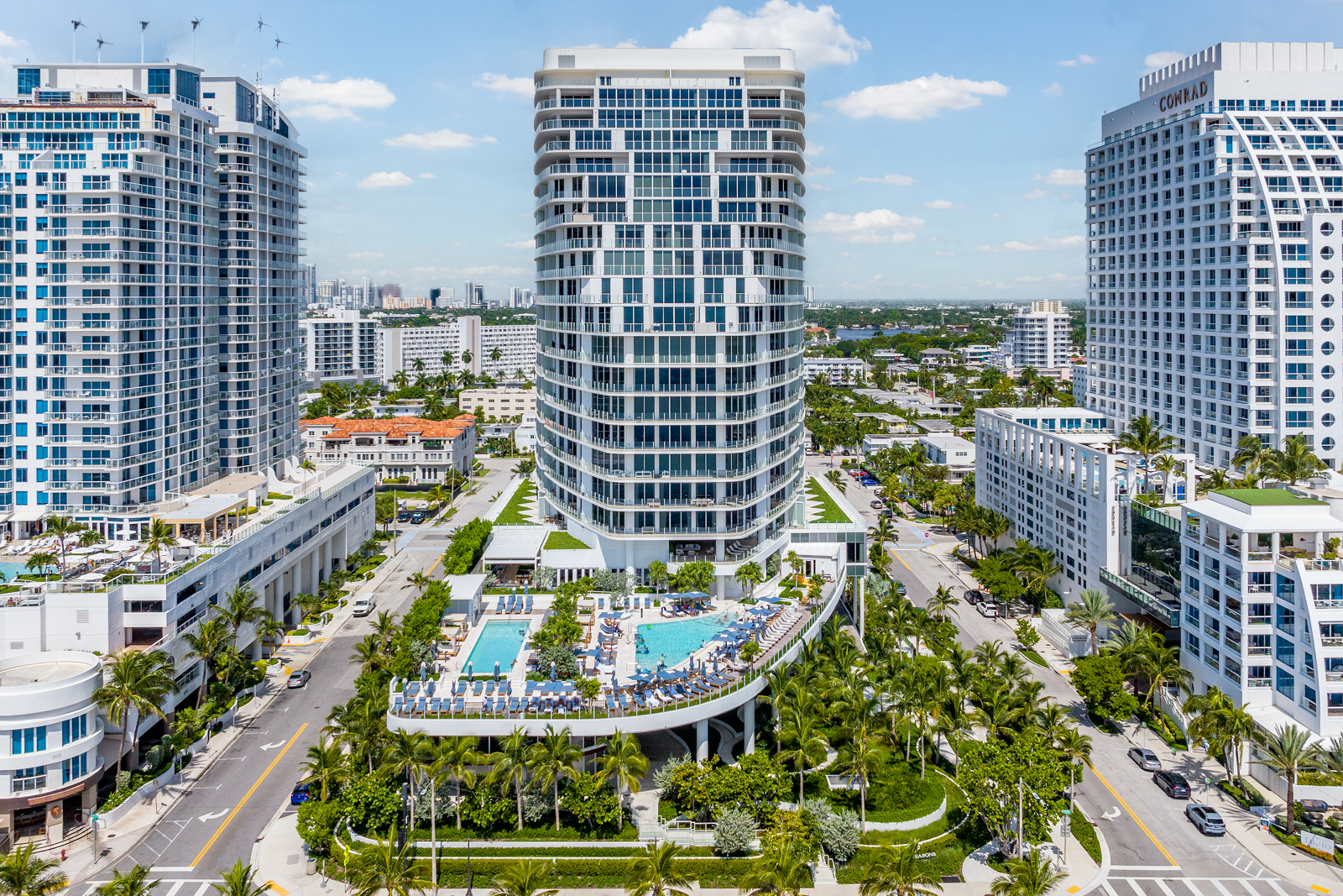 This aerial view showcases a luxury high-rise condominium complex with a prominent central tower and two flanking buildings. The complex features multiple swimming pools, lush landscaping with palm trees, and meticulously maintained grounds. The architecture is modern and sleek, emphasizing the upscale nature of the property and its prime location.