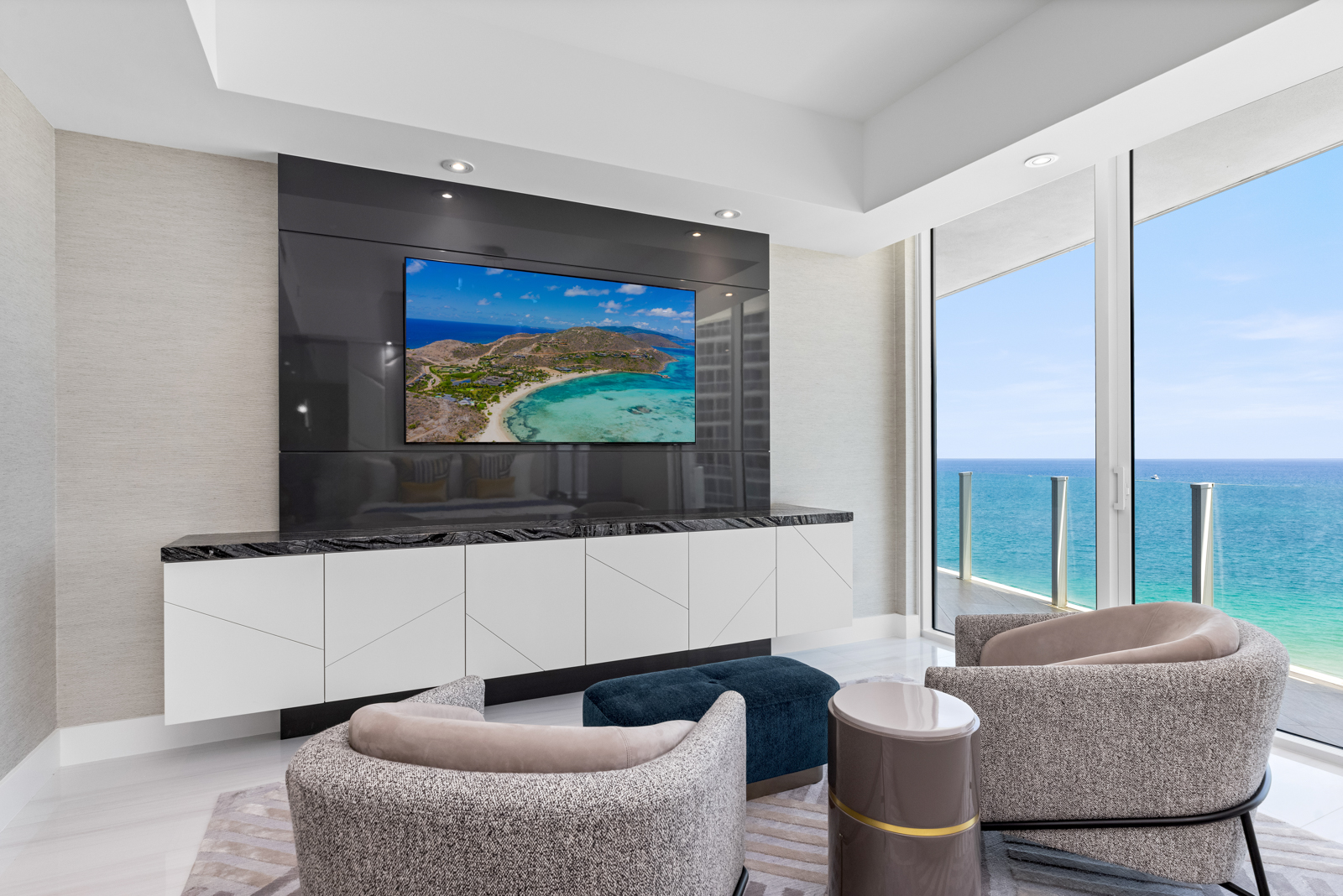 This is a modern living room with a sleek entertainment center featuring a large screen displaying a beach scene. The room is furnished with two stylish armchairs, a small ottoman, and a side table, all arranged on a patterned rug. Large windows offer a stunning ocean view, creating a luxurious and relaxing atmosphere.
