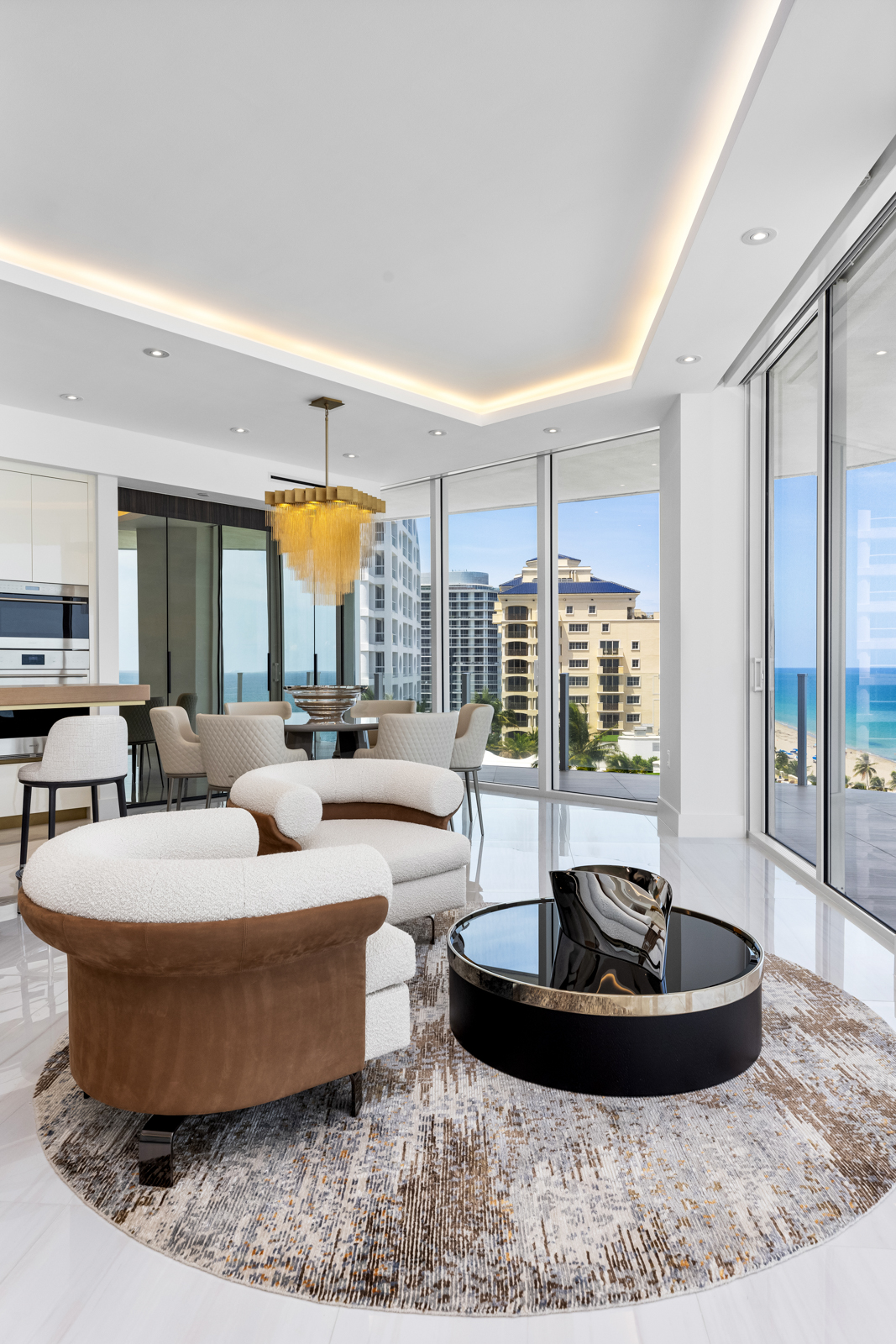 This is a luxurious living room featuring modern furniture, including two stylish armchairs and a round coffee table on a patterned rug. Large windows offer stunning ocean views, enhancing the bright and airy atmosphere. The open-concept design seamlessly integrates with the dining area, creating a sophisticated and inviting space.
