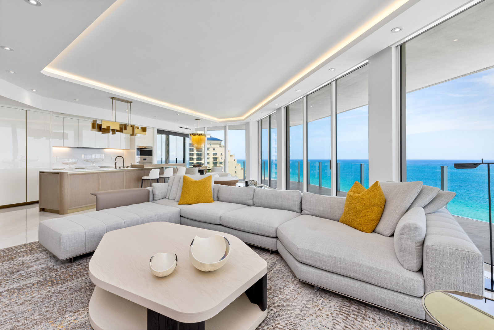 This is a bright and airy living room with floor-to-ceiling windows offering stunning ocean views. The room features a large, sectional sofa with yellow accent pillows, a modern coffee table with decorative bowls, and an open-concept design that flows into the kitchen. The overall aesthetic is luxurious and contemporary, perfect for upscale living.
