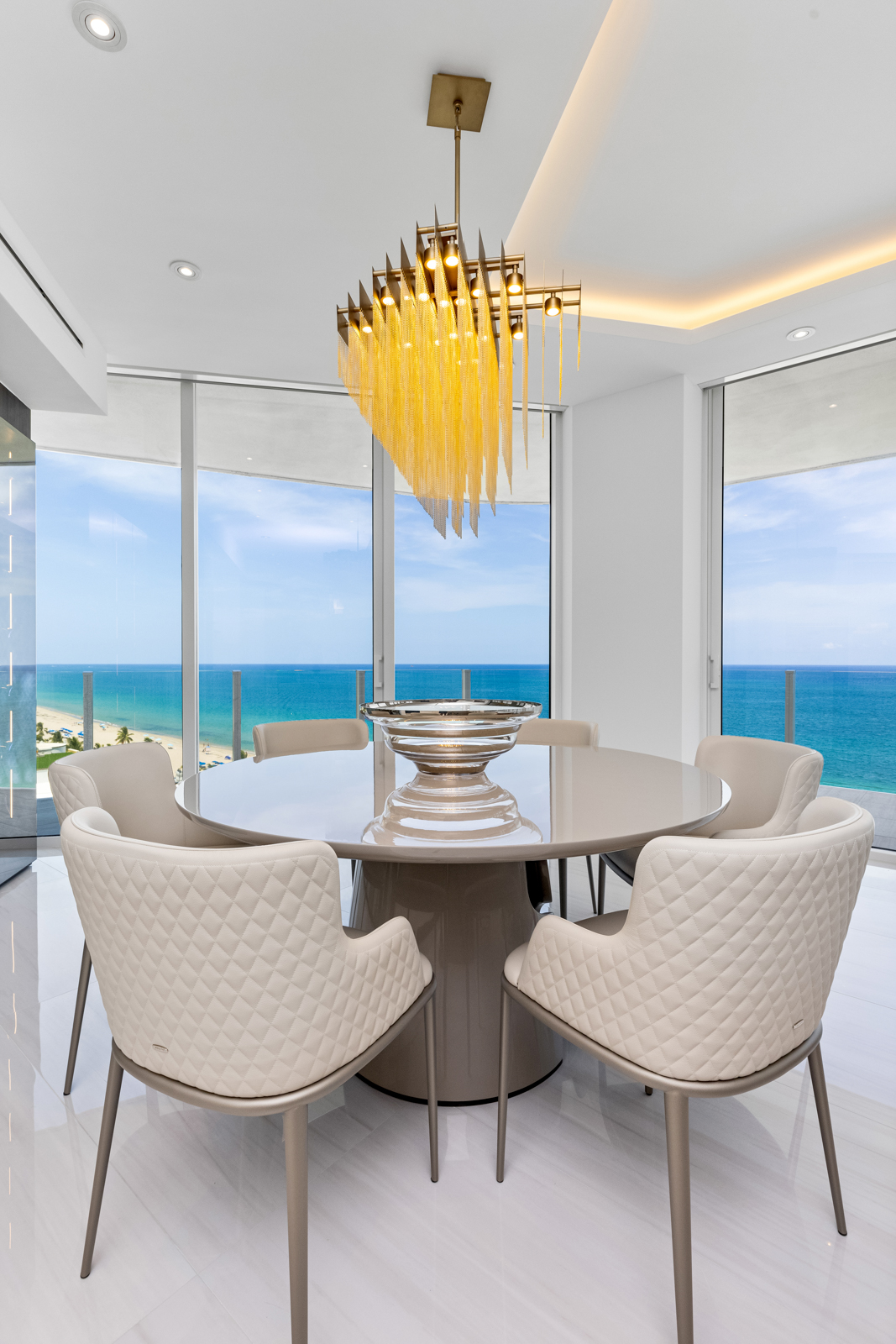 This is a bright and modern dining room featuring a round table with six upholstered chairs. A striking chandelier hangs above the table, and large windows offer stunning ocean views. The room is decorated in neutral tones with white marble flooring, creating a luxurious and airy atmosphere.