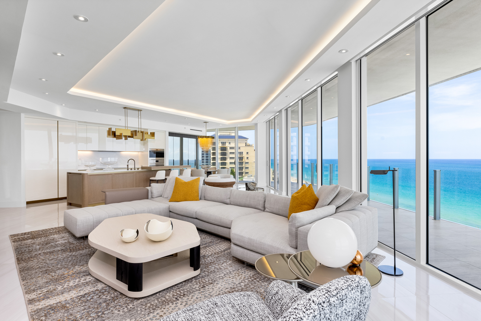 This is a bright and airy living room featuring a large sectional sofa with yellow accent pillows, a modern coffee table with decorative bowls, and floor-to-ceiling windows offering stunning ocean views. The open-concept design seamlessly integrates the living area with the kitchen, creating a spacious and inviting atmosphere. The perspective is from a low angle, emphasizing the room's depth and the expansive view.