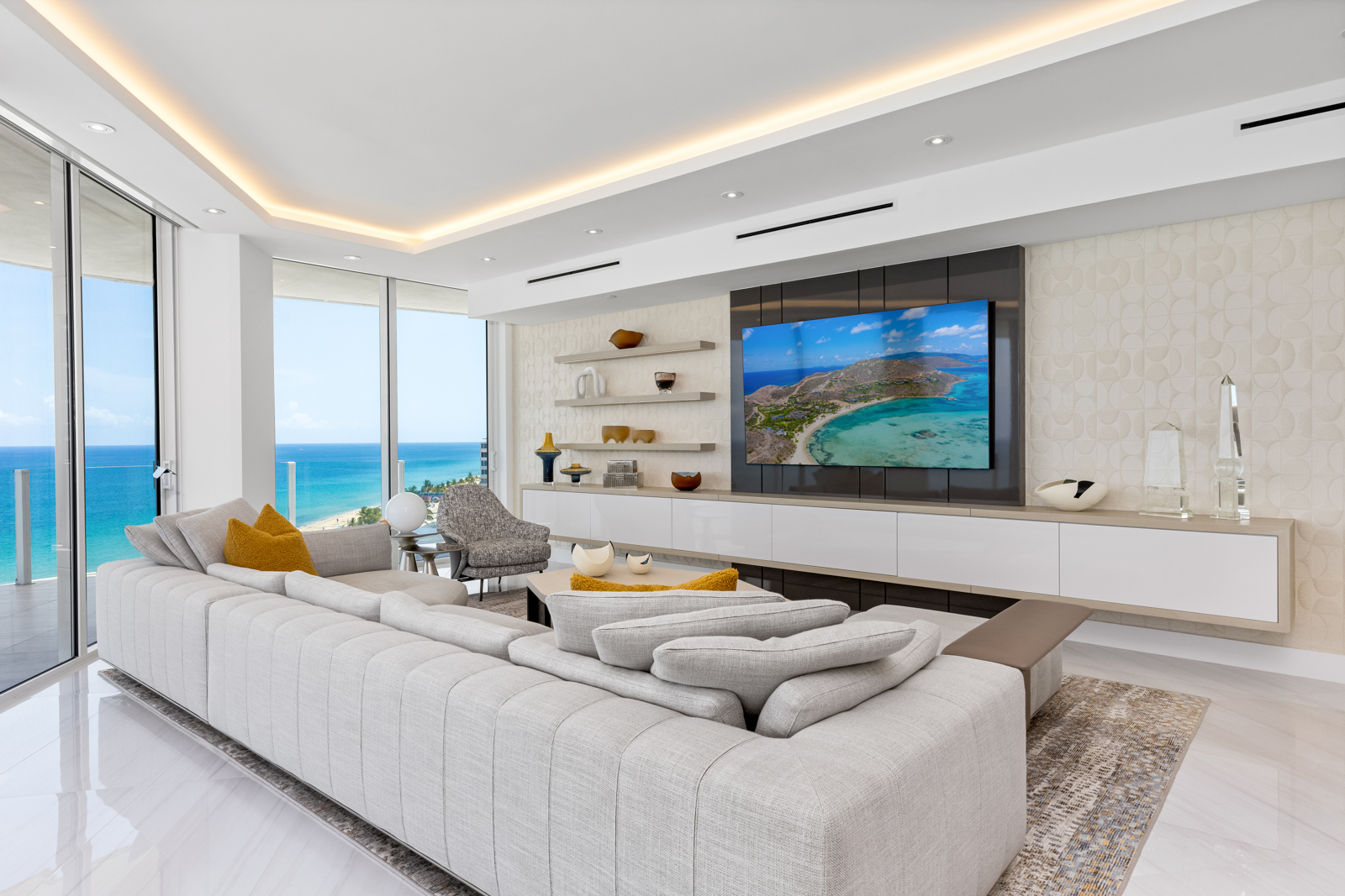This is a bright and modern living room with floor-to-ceiling windows offering ocean views. The room features a large, comfortable sectional sofa, a sleek entertainment center with a large screen TV, and stylish decor. The overall impression is luxurious and inviting, perfect for relaxation and entertaining.