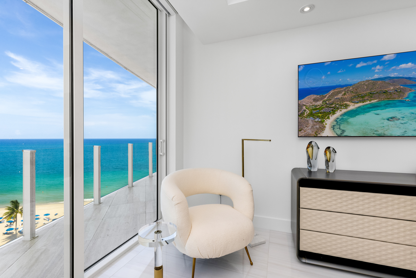 This interior shot showcases a modern living room with a serene ocean view. A stylish, cream-colored armchair sits beside a small, transparent side table, complemented by a sleek dresser with decorative vases and a wall-mounted television displaying a tropical island scene. The room's design emphasizes clean lines and natural light, creating a luxurious and inviting atmosphere.