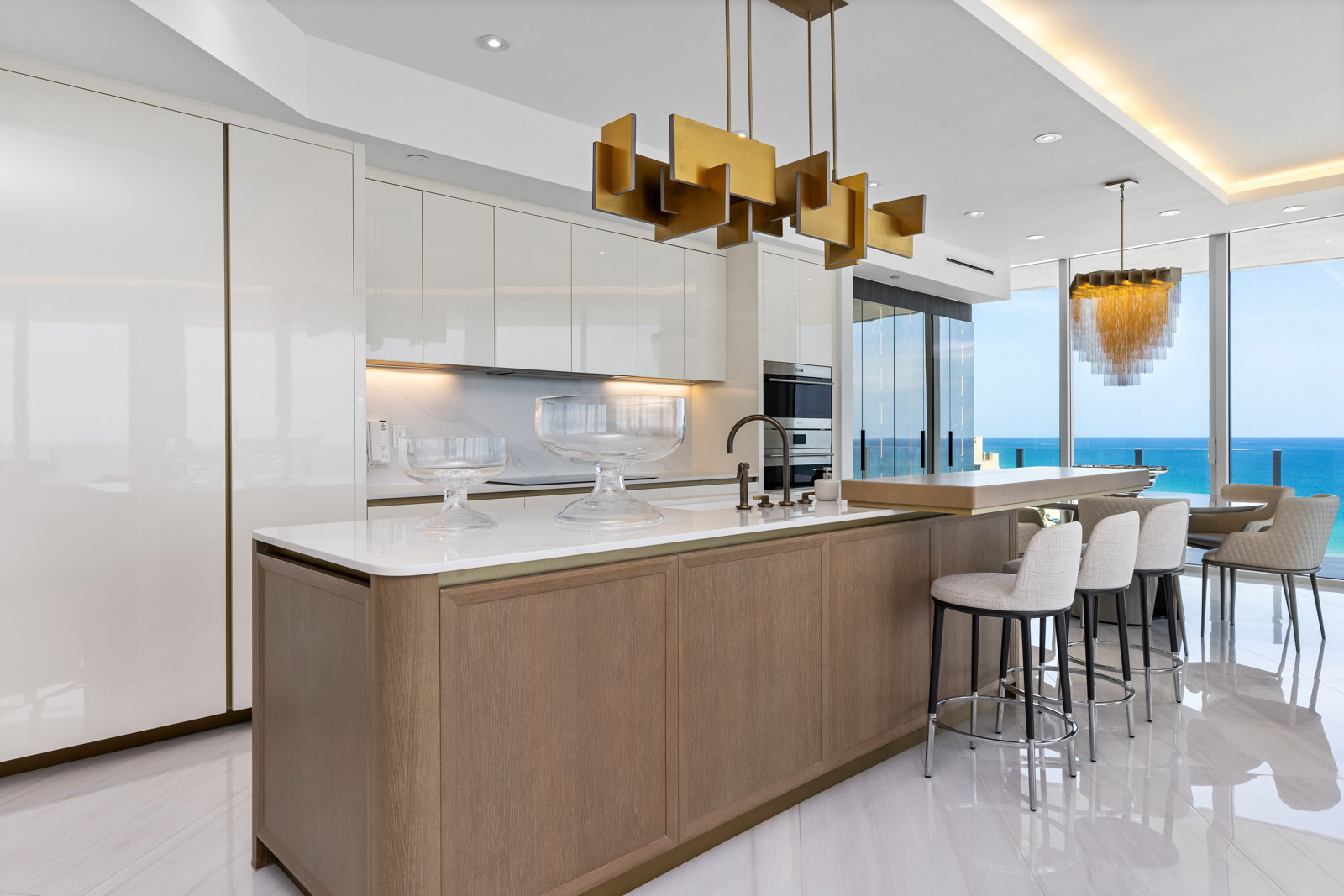 This is a bright and modern kitchen featuring sleek, glossy white cabinetry and a contrasting wood-paneled island with a white countertop. The kitchen is well-lit with both natural light from a large window overlooking the ocean and stylish pendant lighting above the island. The scene is captured from a wide angle, showcasing the open layout and high-end finishes, creating an inviting and luxurious atmosphere.
