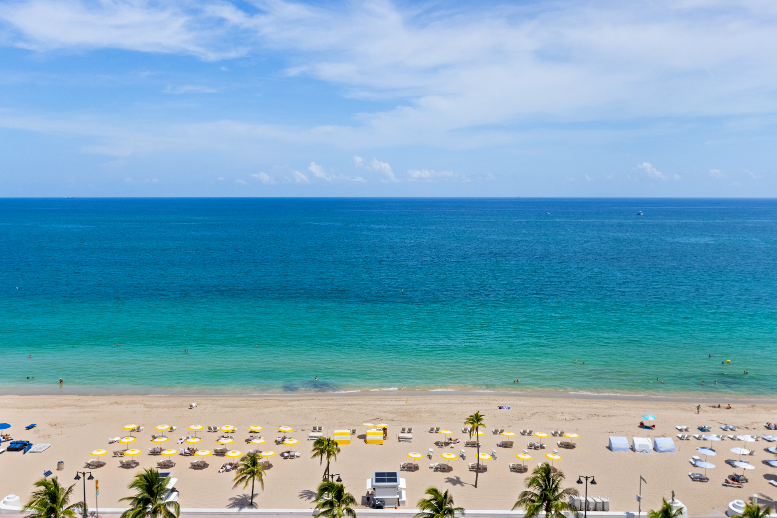 This aerial view showcases a pristine beach with turquoise waters meeting the sandy shore. Rows of yellow and white beach umbrellas and lounge chairs are neatly arranged, suggesting a resort or well-maintained beach area. Palm trees line the edge of the beach, adding a tropical feel to the scene.