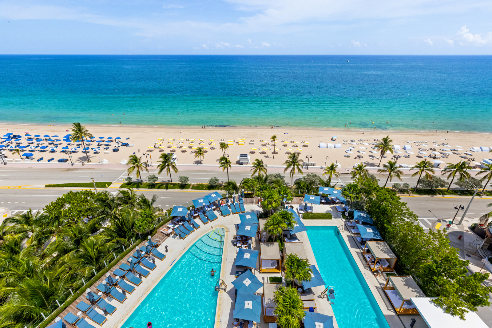This aerial image showcases a luxurious beachfront property, highlighting its expansive pool area with lounge chairs and sun umbrellas. Beyond the pool, a pristine sandy beach meets the turquoise ocean under a bright blue sky. The combination of the pool, beach, and ocean creates an inviting and serene atmosphere, perfect for relaxation and recreation.