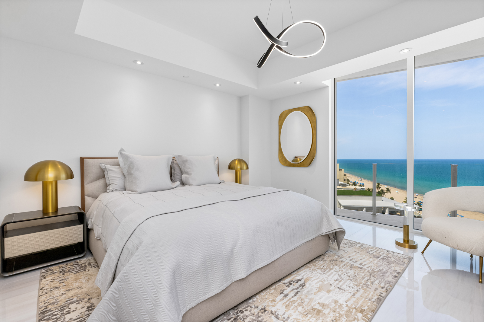 This is a bright and modern primary bedroom featuring a large bed with neutral-toned linens, complemented by contemporary bedside tables and lamps. A striking modern light fixture hangs from the ceiling, and a large window offers a beautiful ocean view, enhancing the room's luxurious and serene atmosphere. The room is decorated with a gold-framed mirror and a stylish chair.