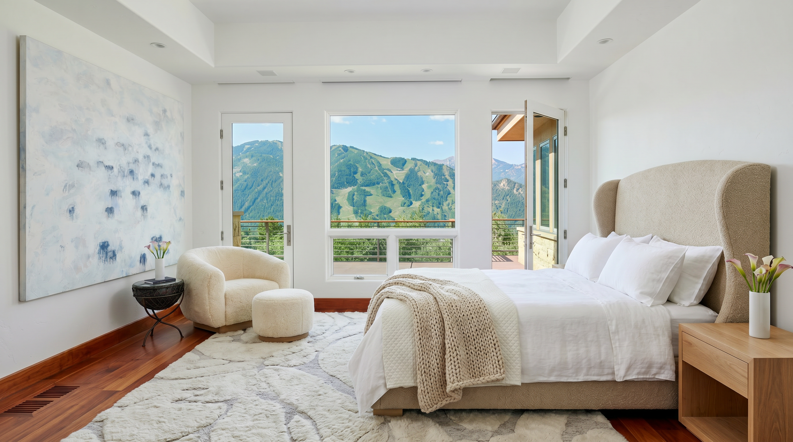 This elegant primary bedroom features a neutral color palette, a plush armchair with an ottoman, and a large abstract painting that adds a touch of artistic flair. The room is bathed in natural light from expansive windows and glass doors that open onto a private balcony, showcasing breathtaking mountain views. The combination of hardwood floors, a textured area rug, and high-end finishes creates a serene and luxurious atmosphere.