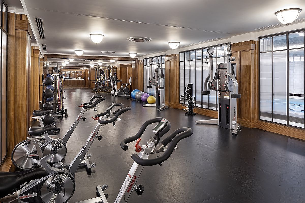 This is a well-equipped home gym featuring several Keiser spin bikes in the foreground, along with a rack of medicine balls and various weight machines in the background. Large windows provide natural light and a view of a pool, while wood paneling adds a touch of elegance to the space. The black flooring gives the room a sleek and modern feel.