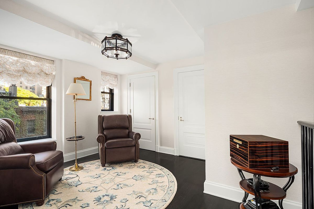 This interior shot showcases a cozy living room area with two brown leather armchairs positioned on a round rug with a floral pattern. A floor lamp with a small side table stands between the chairs, and natural light streams in through a window with decorative valances. The room features dark hardwood floors and neutral-toned walls, creating a warm and inviting atmosphere.