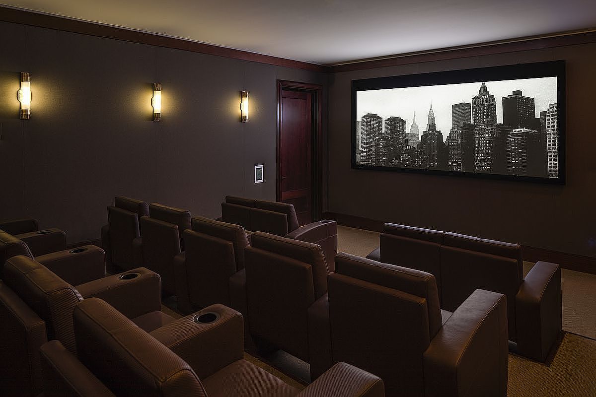 This is an interior shot of a home theater. The room features multiple rows of leather seating with cup holders, facing a large screen displaying a black and white cityscape. Wall sconces provide ambient lighting, and a dark wood door is visible on the back wall, creating a luxurious and immersive viewing experience.