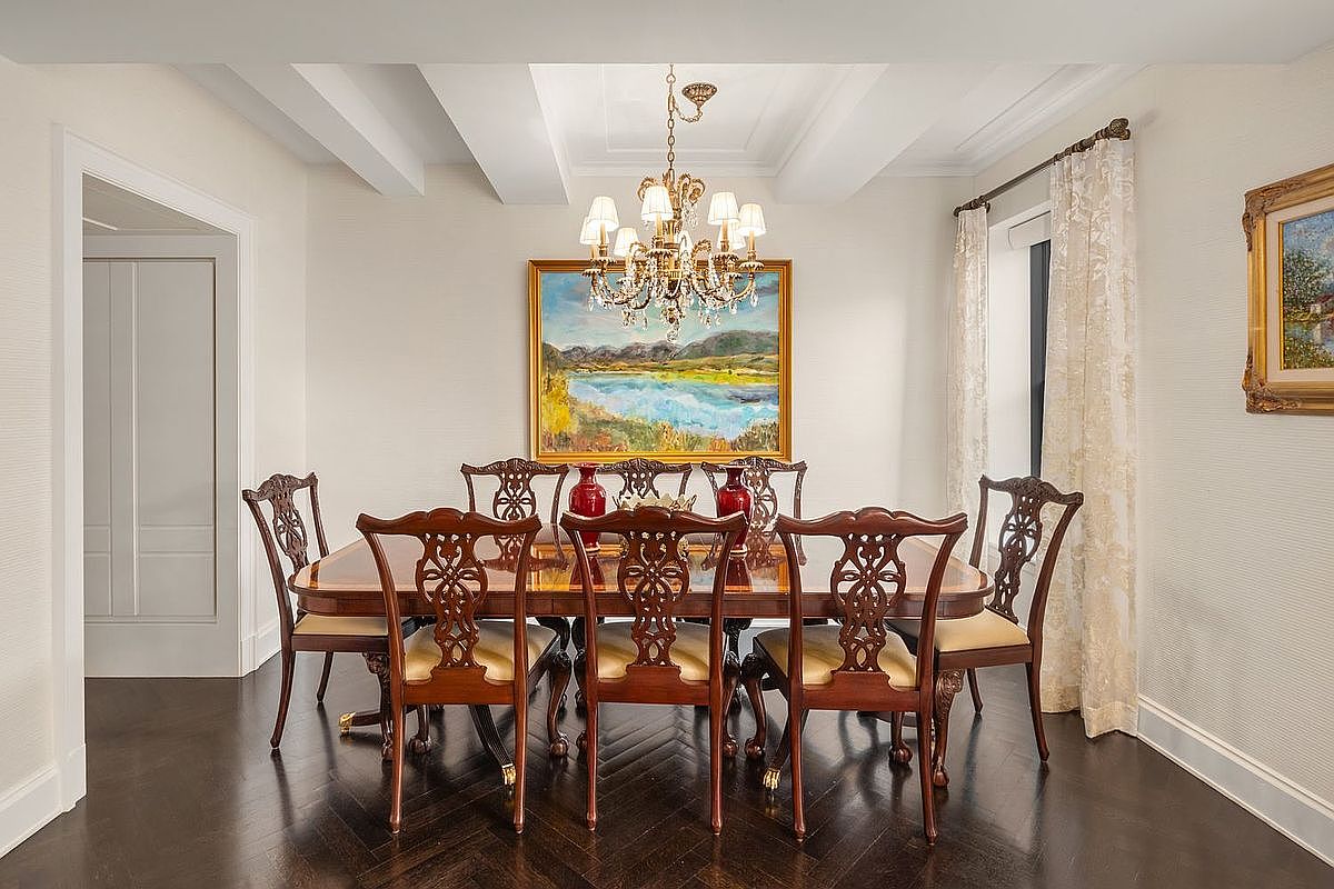 This is an interior shot of a formal dining room. The room features a large wooden dining table with six chairs, a chandelier hanging above the table, and a painting on the wall. The walls are textured, and there is a doorway leading to another room, creating a sense of spaciousness and elegance.