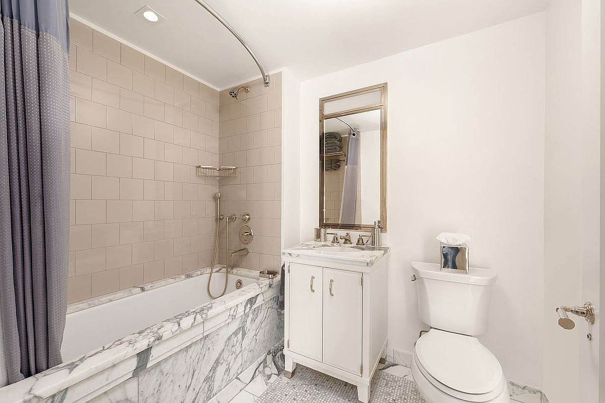 This bathroom features a bathtub with a marble surround and a gray shower curtain. A white vanity with a marble countertop and a framed mirror sits next to a toilet with a tissue box on top. The floor is tiled with small mosaic tiles, and the walls are painted a neutral color, creating a clean and classic look.