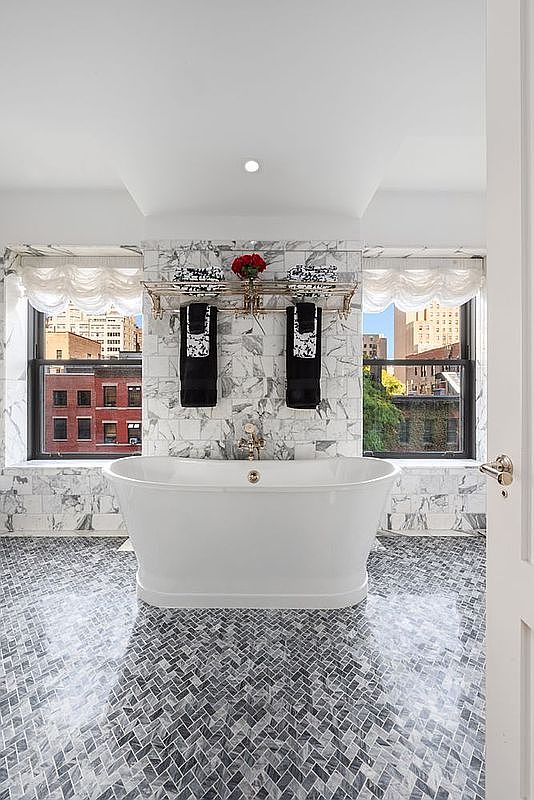 This is a luxurious primary bathroom featuring a freestanding white bathtub as the focal point, set against a backdrop of marble-tiled walls and a herringbone-patterned floor. Natural light floods the space through two large windows, complemented by elegant window treatments and a decorative towel rack with black towels. The overall impression is one of sophistication and relaxation.
