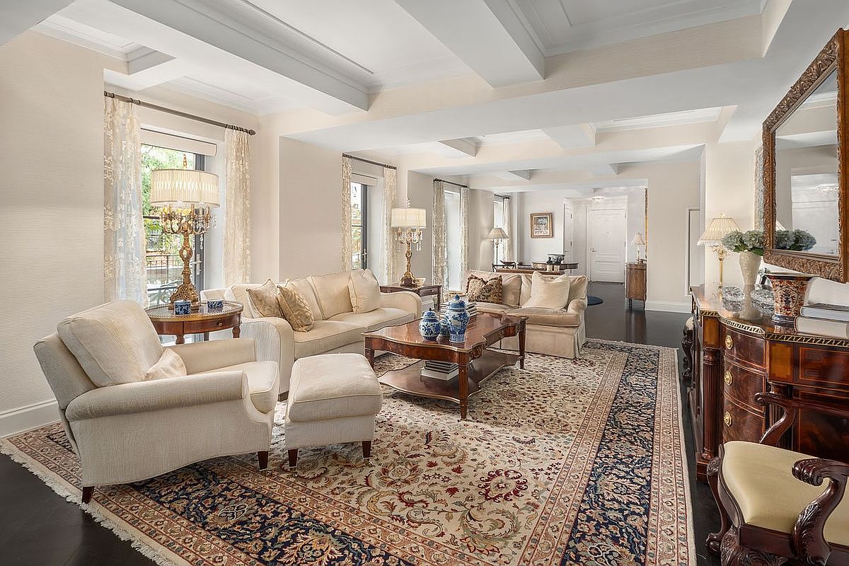 This is a well-appointed living room featuring two sofas, an armchair with an ottoman, and a dark wood coffee table atop an ornate area rug. The room is illuminated by table lamps and natural light filtering through the windows, which are adorned with sheer curtains. The coffered ceiling adds architectural interest, and a dark wood console with a large mirror completes the elegant and traditional aesthetic.
