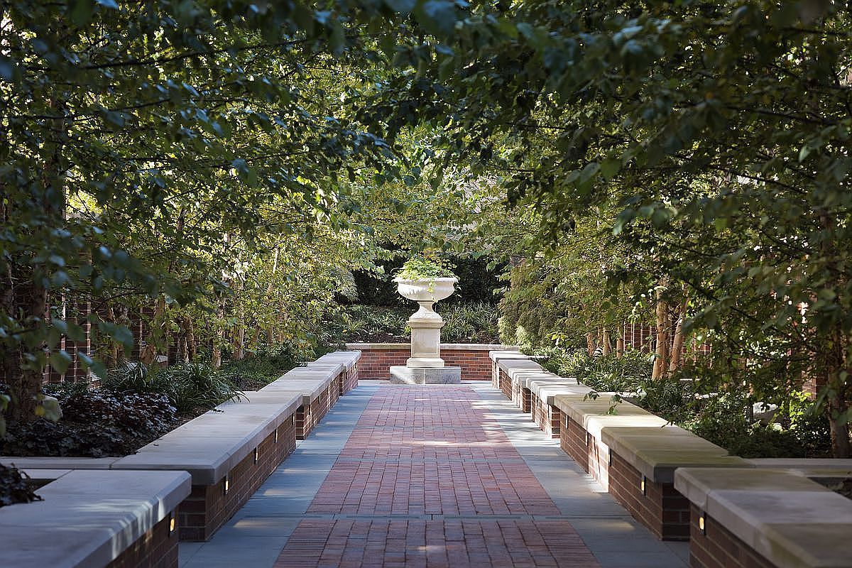 This image showcases a beautifully landscaped garden path leading to a decorative urn. The path is constructed of red brick and bordered by stone walls and lush greenery, creating a serene and inviting atmosphere. Mature trees line the path, providing shade and enhancing the garden's tranquil ambiance.
