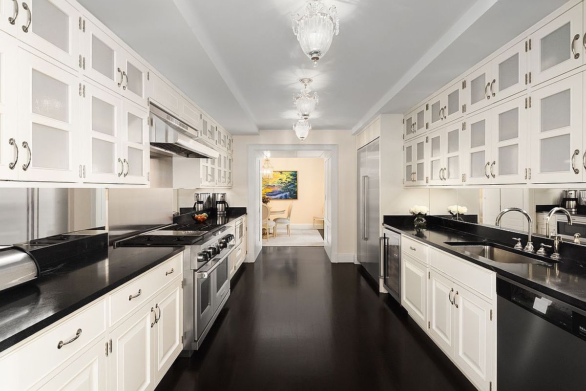 This is a galley kitchen with dark hardwood floors and white cabinets. The countertops are black, and stainless steel appliances are visible. The kitchen is well-lit with decorative light fixtures and leads into another room through a doorway.